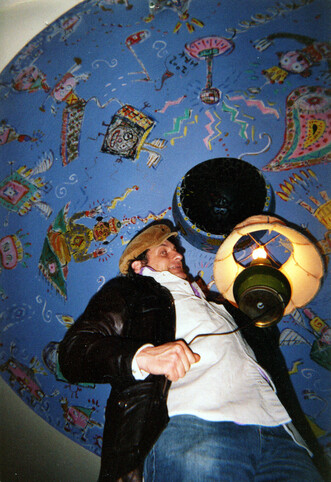 Hamish Kilgour at WFMU sometime around 1999 holding a hanging lamp underneath the dome he painted. The background color of the dome is blue and there are numerous whimsical characters painted on the dome.