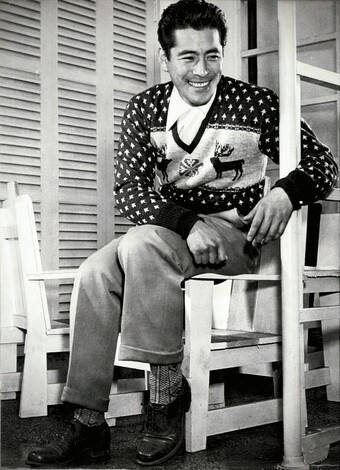 A young Tohsiro Mifune is sitting in a chair and casually laughing, looking quite handsome in a Christmas sweater with two deer and a snowflake on it.