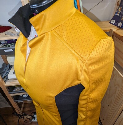A yellow and black minidress uniform from Star Trek: strange New Worlds sits partially constructed on a dress form. The shoulder and the outer sleeve have been laser etched with the repeating star pattern from the show, which for the record took over five hours and came out looking brilliant. The front is pinned closed because it's unfinished, and several seams need ironing and topstitching.