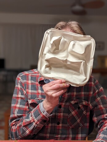 A person holding a mask made from a mangled takeout food box in front of his face