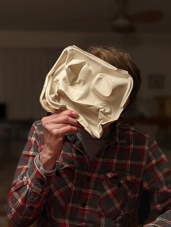 A person holding mask made from a mangled takeout food box in front of his face