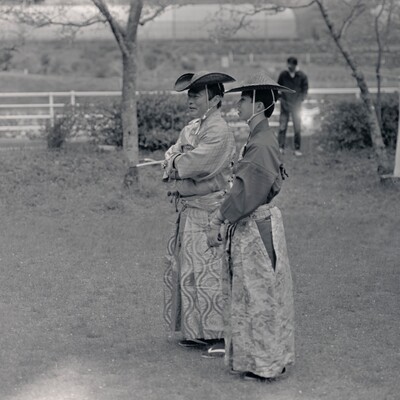 Black and white photograph of two men—possibly father and son— dressed in period Japanese attire, talking to one another at a mounted archery competition.